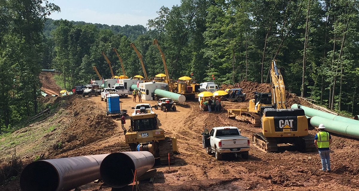 A worksite involves cranes, excavators, company vehicles, and large pipeline segments on a cleared right-of-way in