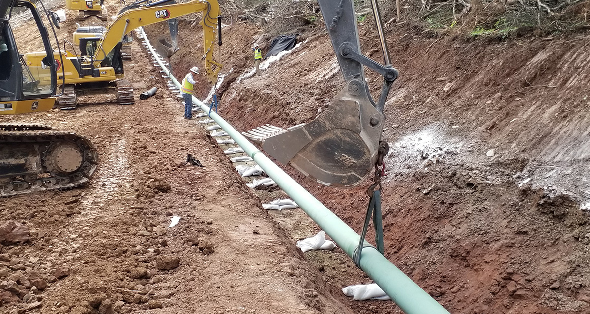 Excavators are lowering an assembled pipeline into a trench at a construction site.